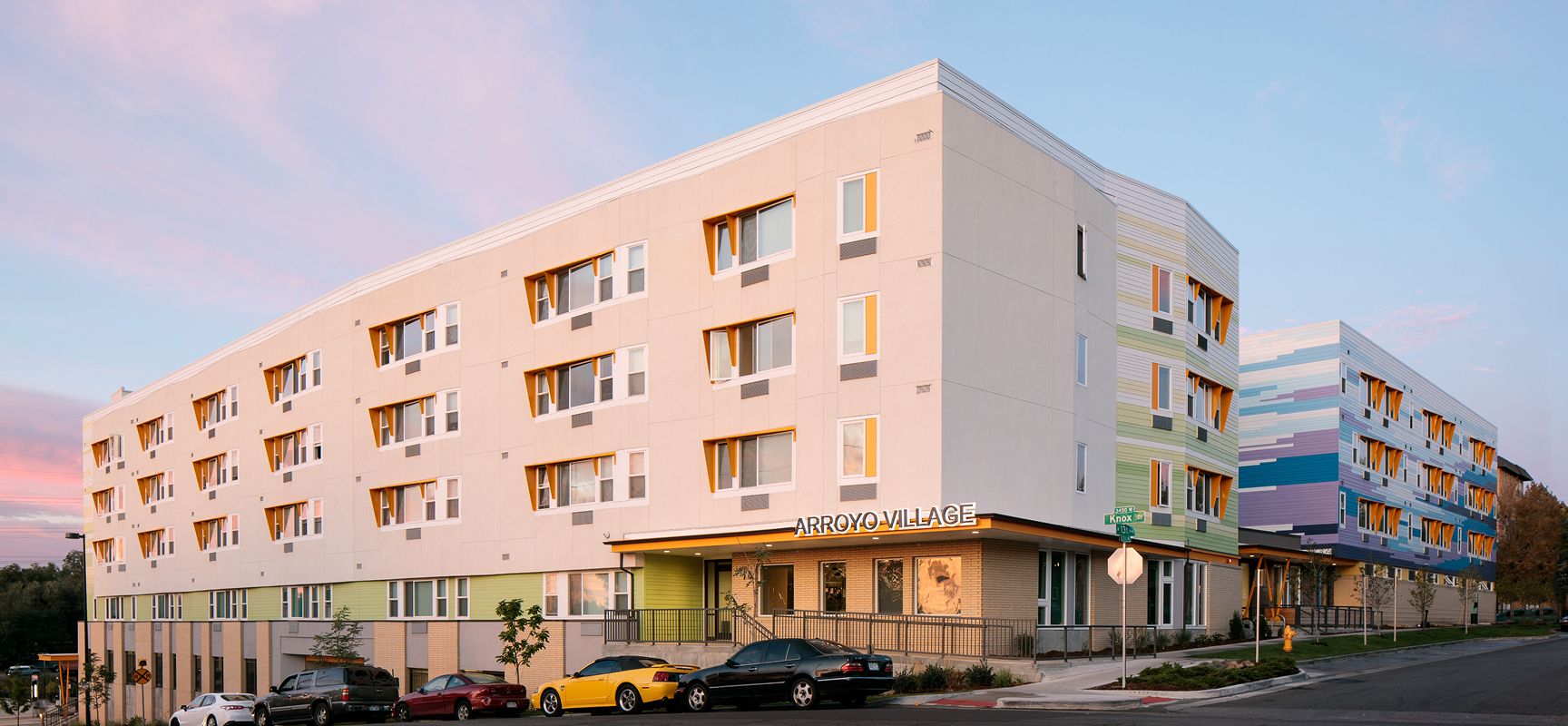 Arroyo Village apartment building at sunset with colorful design and parked cars.