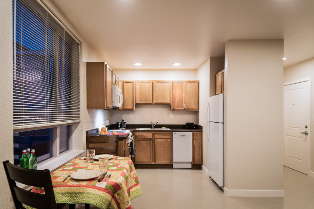 Small kitchen with wooden cabinets, dining table with colorful cloth, and window blinds.