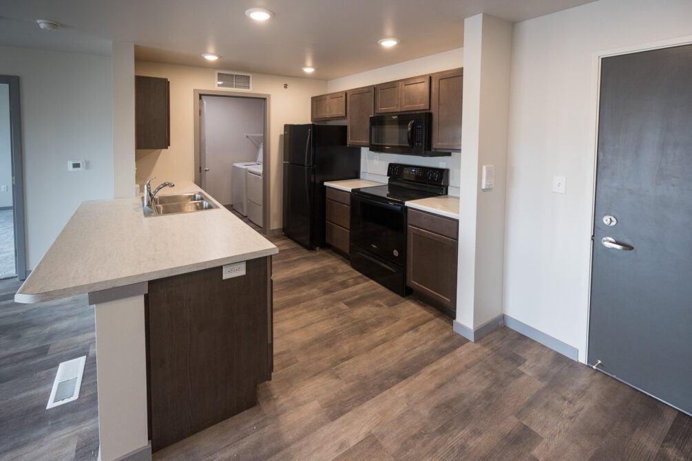 Modern kitchen with dark wooden cabinets, stainless steel sink, and black appliances. Laundry room visible in the background.