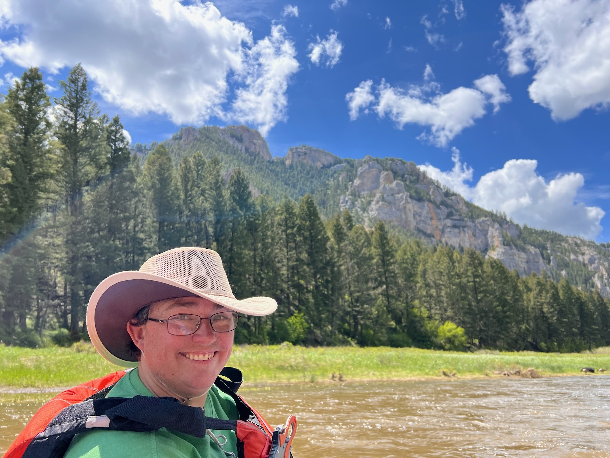 Ashley smiling with a wide-brimmed hat, river and forested mountains in background
