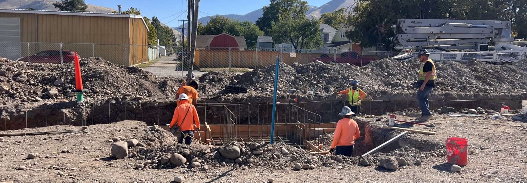 Construction workers wearing safety gear at an excavation site with machinery and mountains in the background