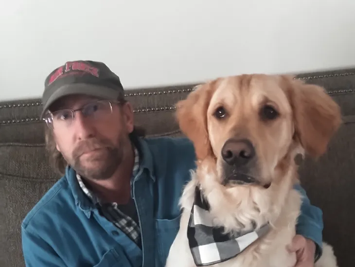 Brett Jackson sitting on a couch with a Golden Retriever wearing a checkered bandana