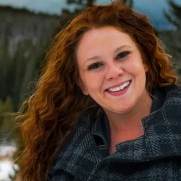 Cass smiling outdoors, wearing a checkered coat, with a forest background