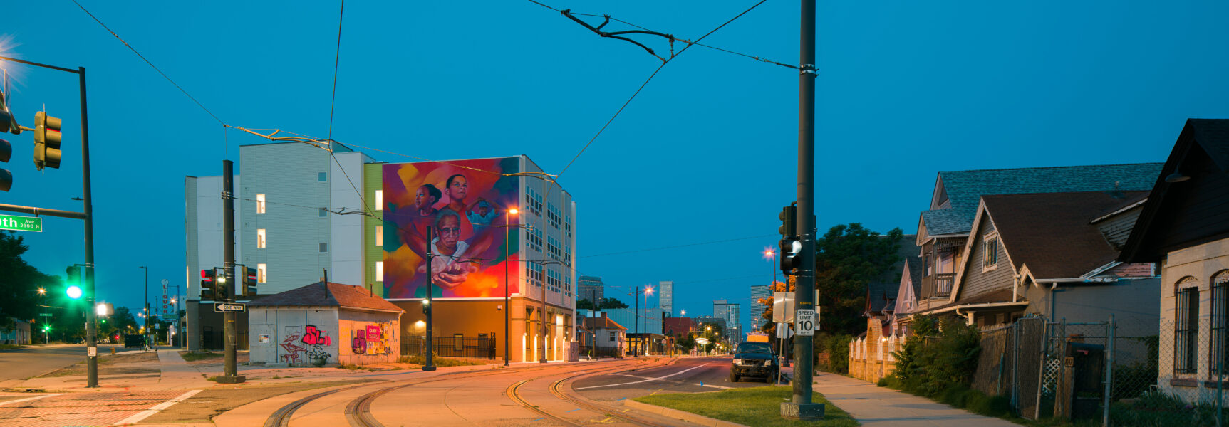 Street view at dusk with vibrant mural on building, depicting three figures, located at the intersection of 29th and Welton.