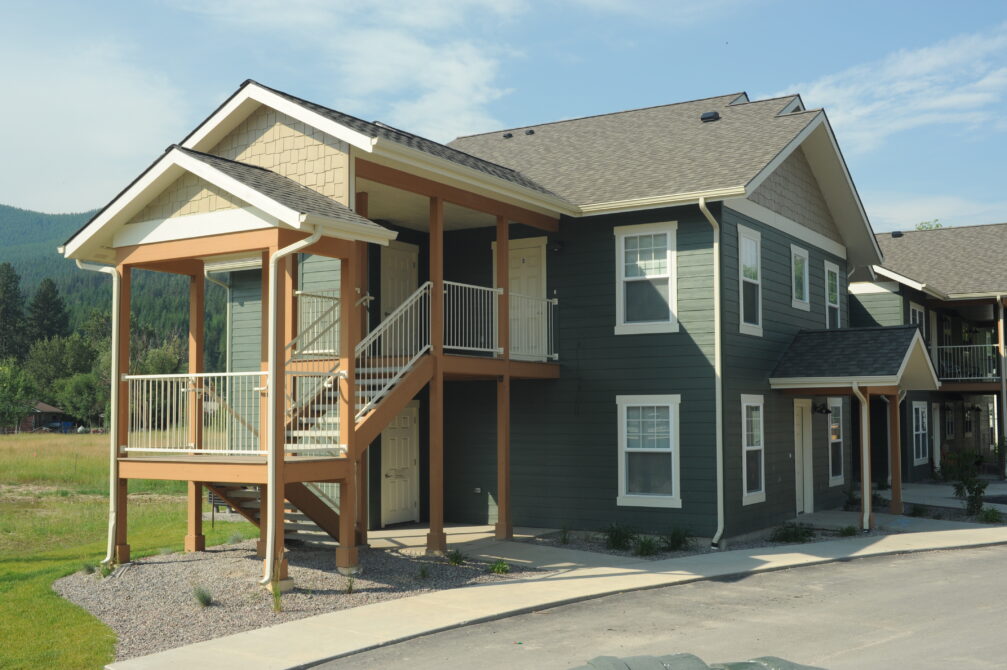 Two-story green townhouse with white trim, outdoor stairs, and porch; set in a grassy, mountainous area.