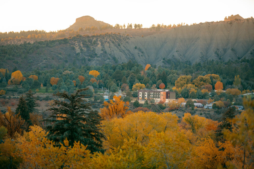 Autumnal landscape with colorful trees, a building, and a mountainous backdrop.