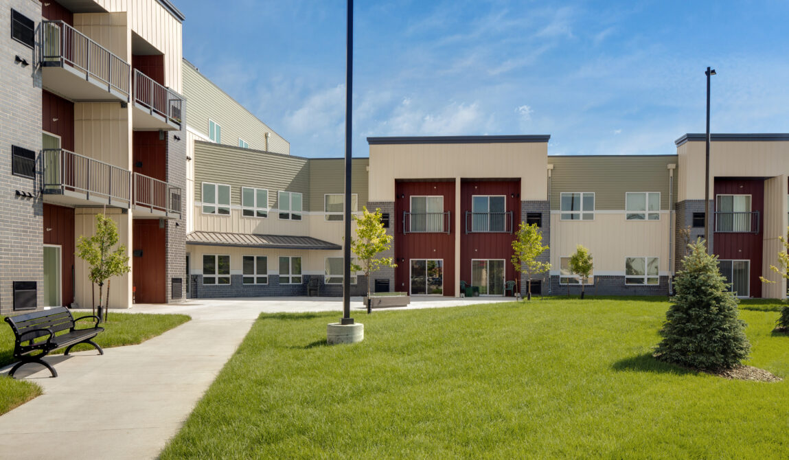 Modern multi-story building with balconies, surrounded by green lawn and trees.