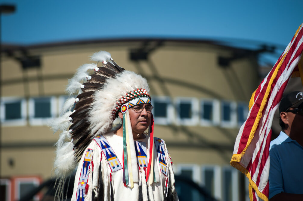 Person in traditional attire with a feathered headdress, holding a flag, standing outdoors