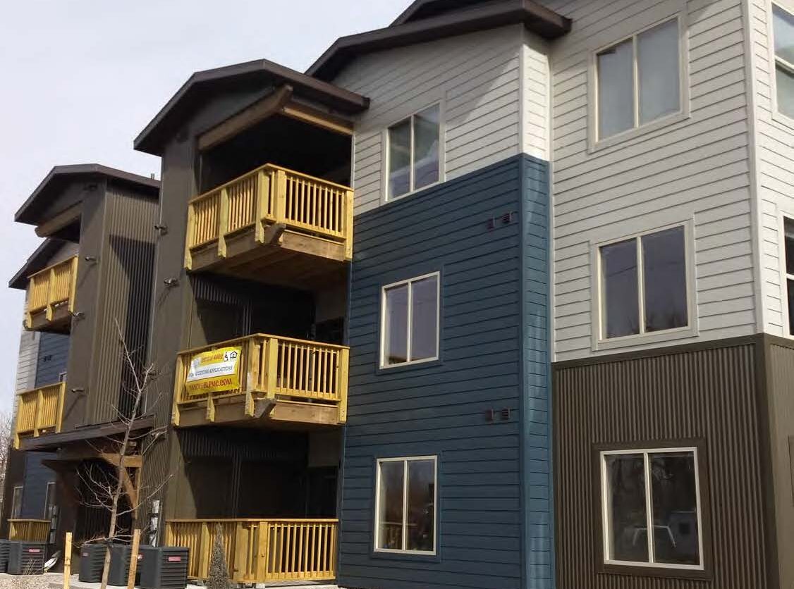 Modern three-story apartment building with wooden balconies and a sign reading "Emerald Construction" on the middle balcony.