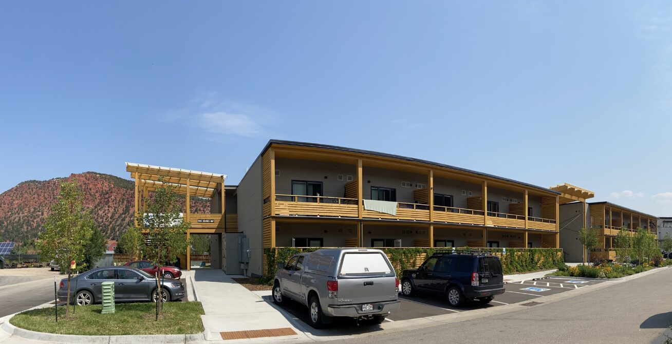 Two-story yellow building with parked cars, surrounded by trees, and a mountain backdrop.
