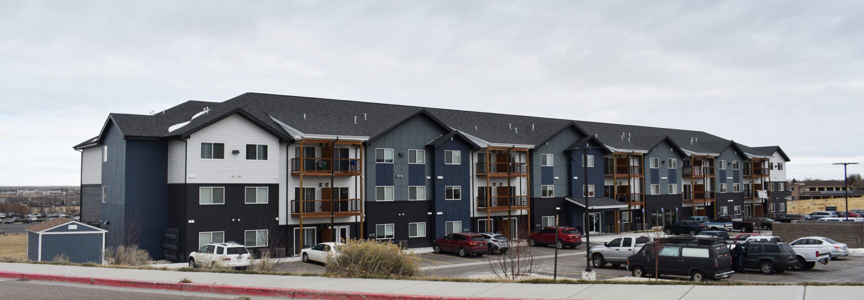 Three-story apartment building with dark blue and white exterior, balconies, and parked cars.