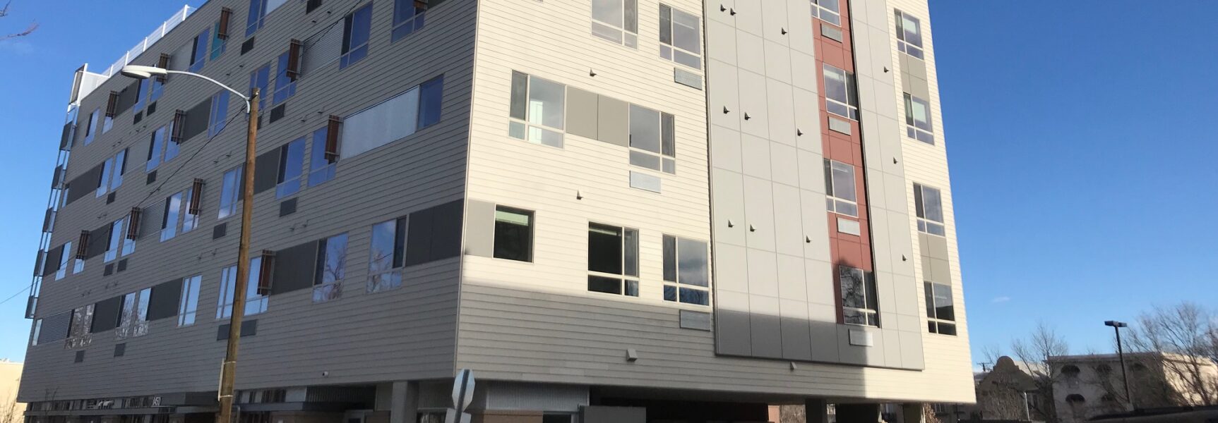 Modern multi-story building with varied window sizes, light panels, and red accents against a clear blue sky