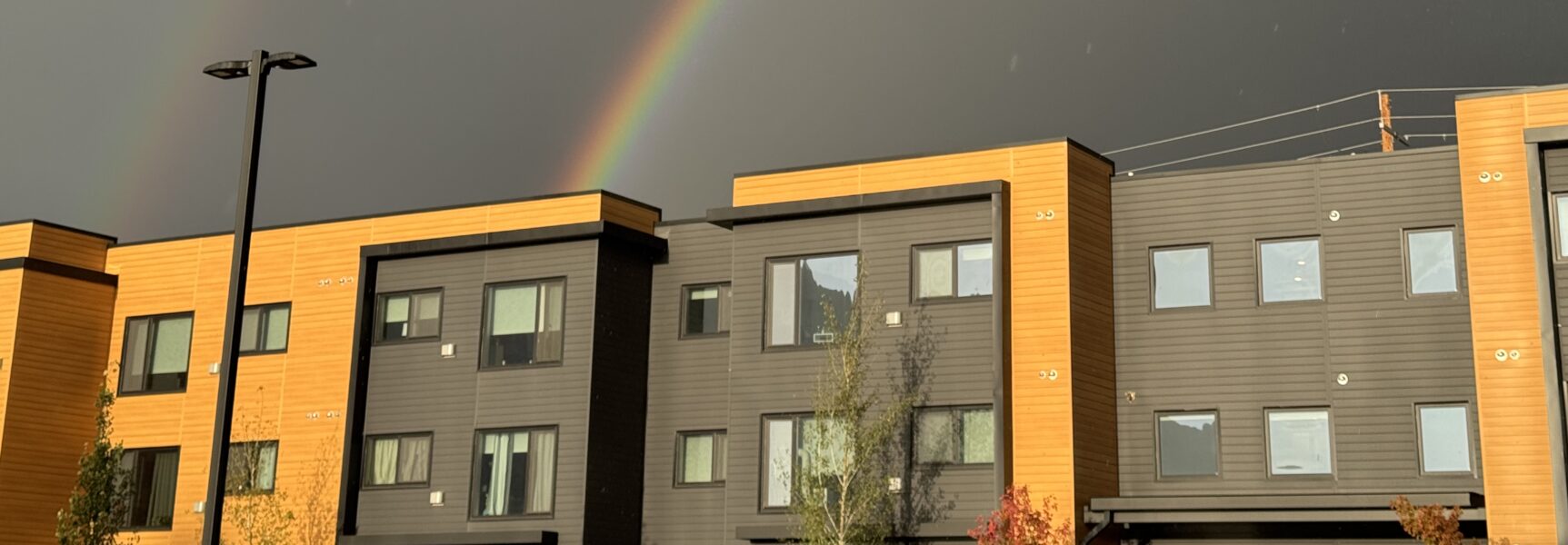 Modern apartment building with wood paneling under a vibrant double rainbow in cloudy sky