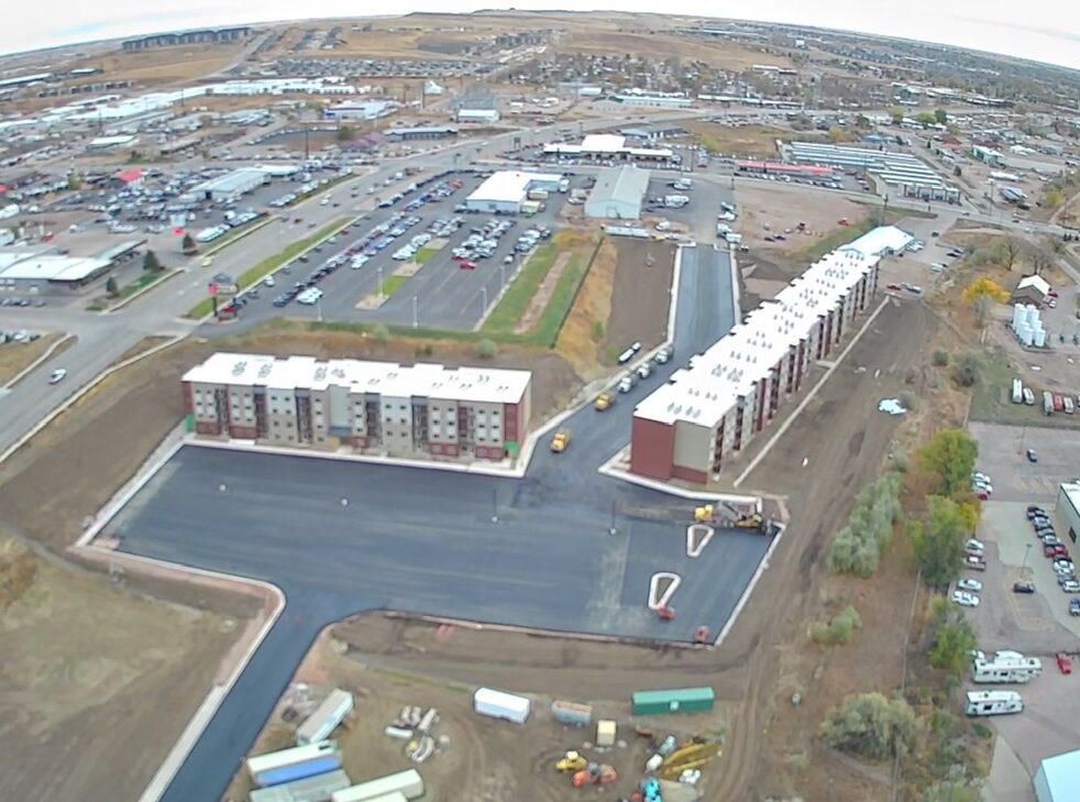 Aerial view of new apartment complex under construction with parking lot, surrounded by roads and commercial area.