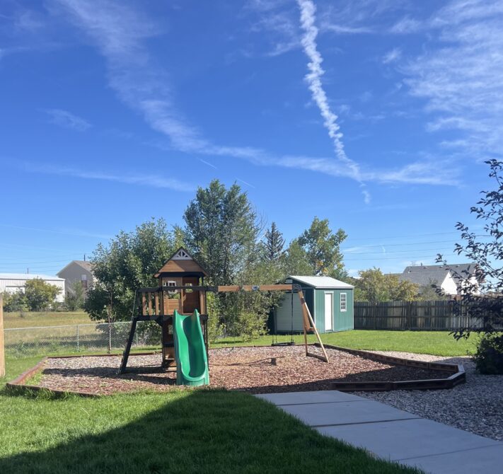Playground set with green slide, wooden swing, and green shed in backyard. Clear blue sky with contrails.