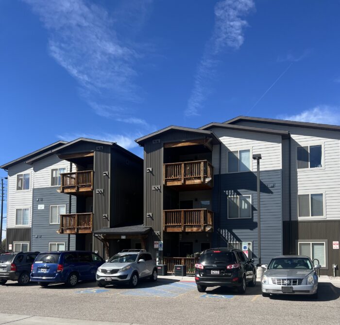 Two apartment buildings with balconies, parked cars, and a clear blue sky