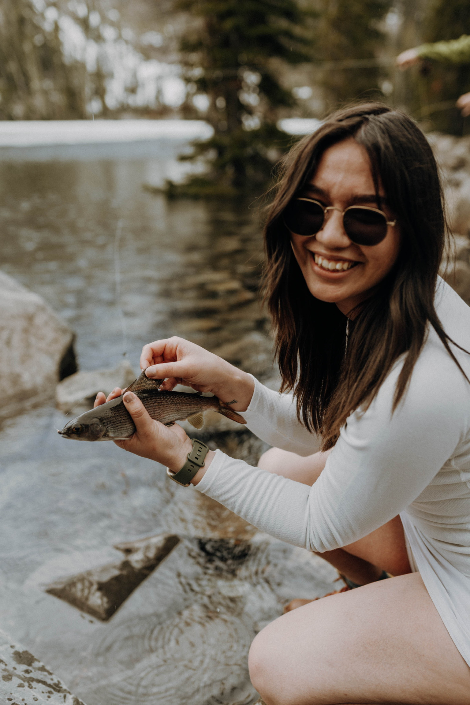 Liz smiling while holding a small fish by a riverside