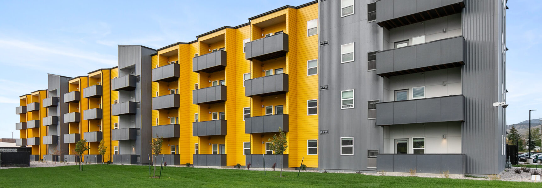 Modern apartment building with vibrant yellow accents and gray balconies, surrounded by green lawn.