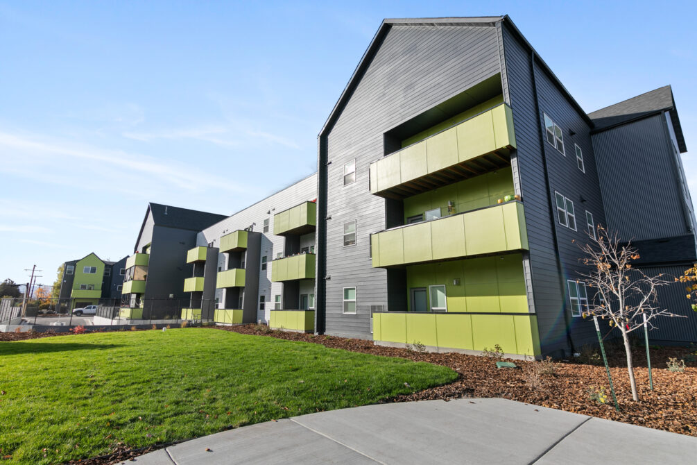Modern apartment building with green and gray exterior, surrounded by grass and trees.