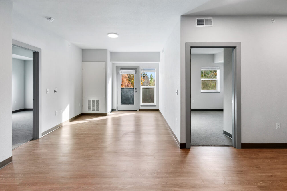 Empty, bright apartment interior with light wood flooring and two doorways leading to carpeted rooms