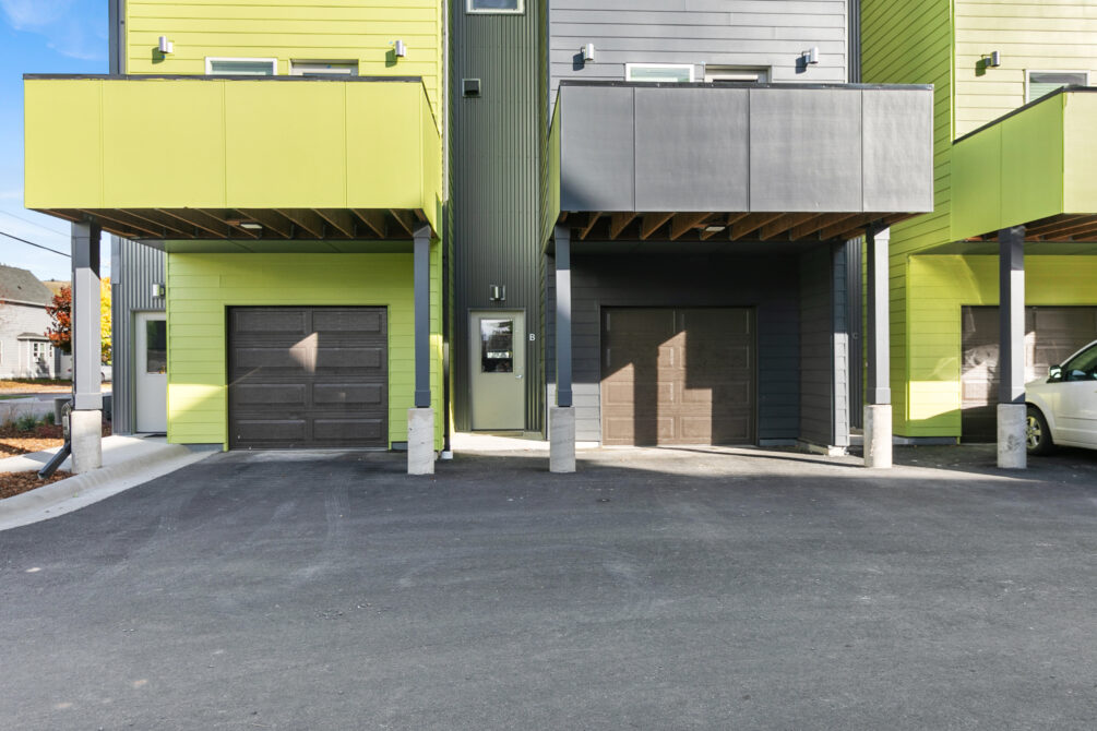 Two modern townhouses with garages, lime green and dark gray facades, brightly lit by sunlight.
