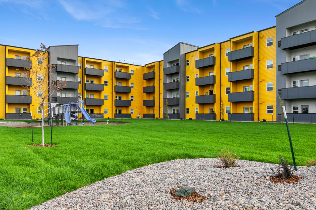Yellow apartment buildings with balconies, a playground, and a grassy courtyard under a blue sky.
