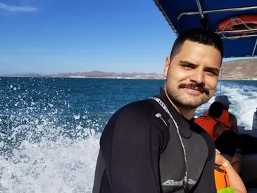 Mitch on a boat at sea, smiling, wearing a wetsuit with a backdrop of splashing water and distant hills.