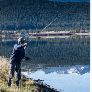 Nate fishing by a lake surrounded by forested hills under a clear blue sky.