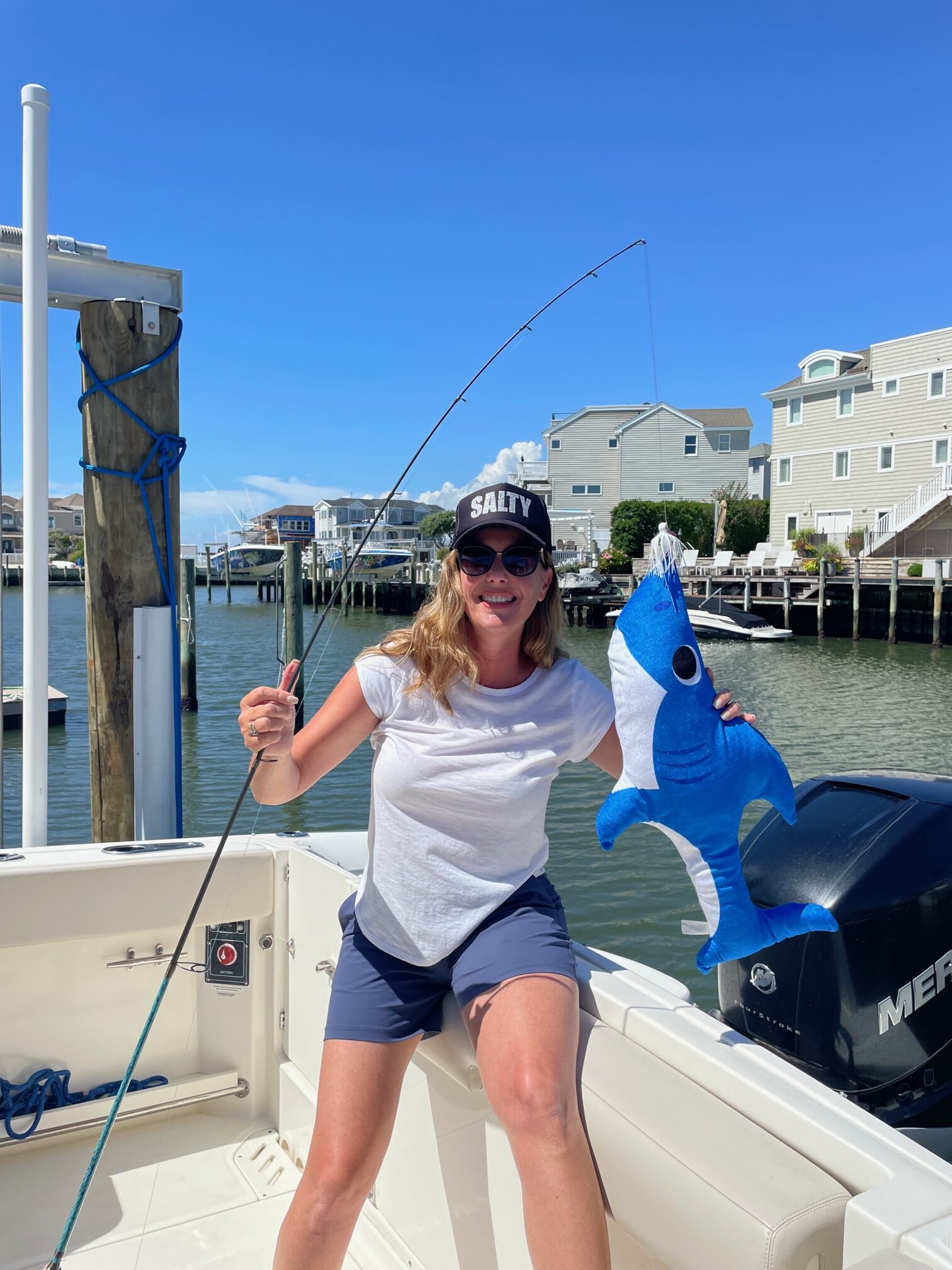 Woman on boat holding fishing rod and blue stuffed fish, wearing "Salty" hat, smiling.