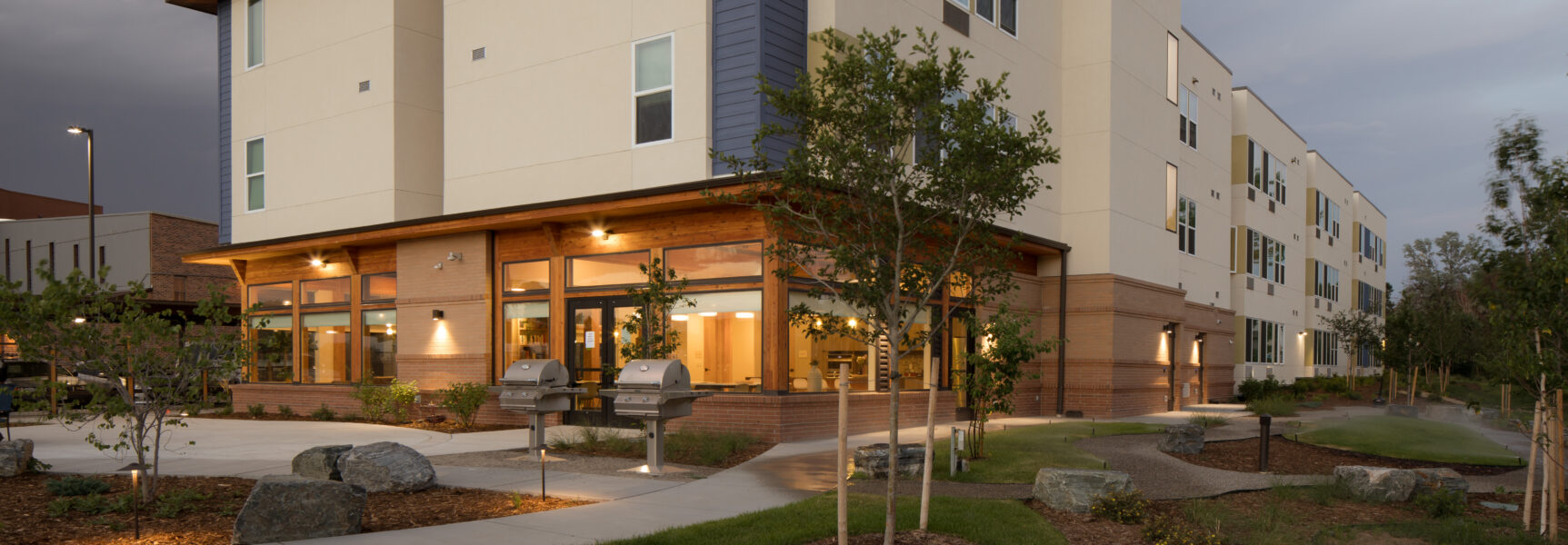 Modern multi-story building exterior with brick and beige walls, large windows, and outdoor patio illuminated at dusk.