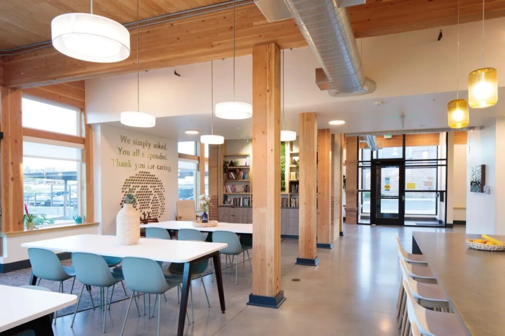 Modern communal space with wooden beams, blue chairs, large tables, bookshelves, and a wall sign reading, "We simply asked. You all responded. Thank you for caring."
