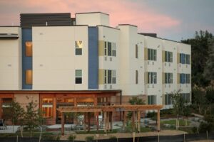 Modern multi-story building with large windows, surrounded by landscaping and a wooden pergola.
