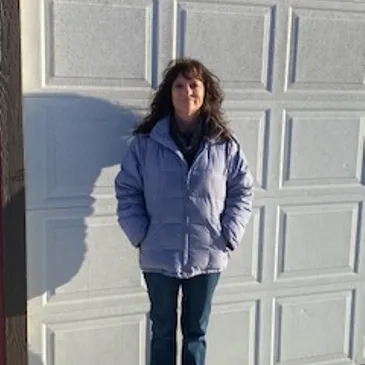 Rebecca stands outside, wearing a light blue jacket, against a garage door.