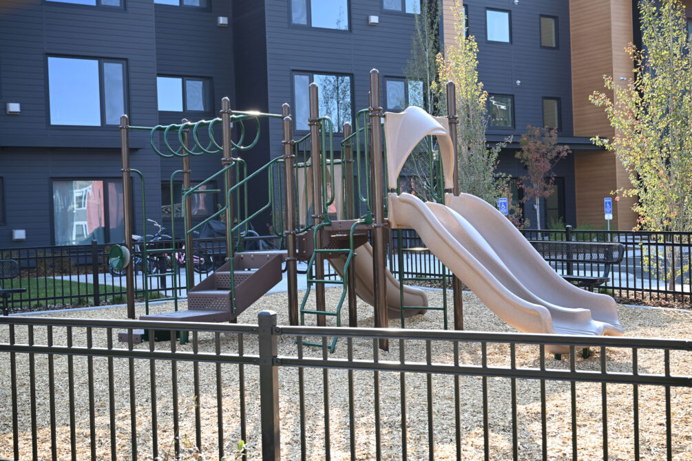 Playground with slides and climbing equipment, surrounded by a fence, near modern buildings