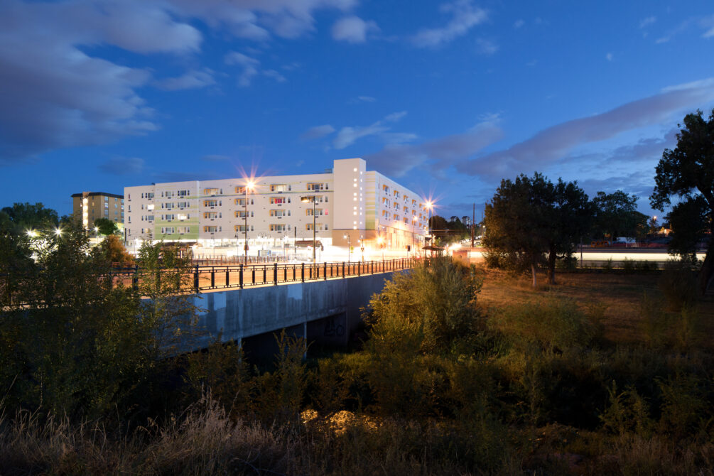 Modern apartment building illuminated at night with trees and a bridge in foreground
