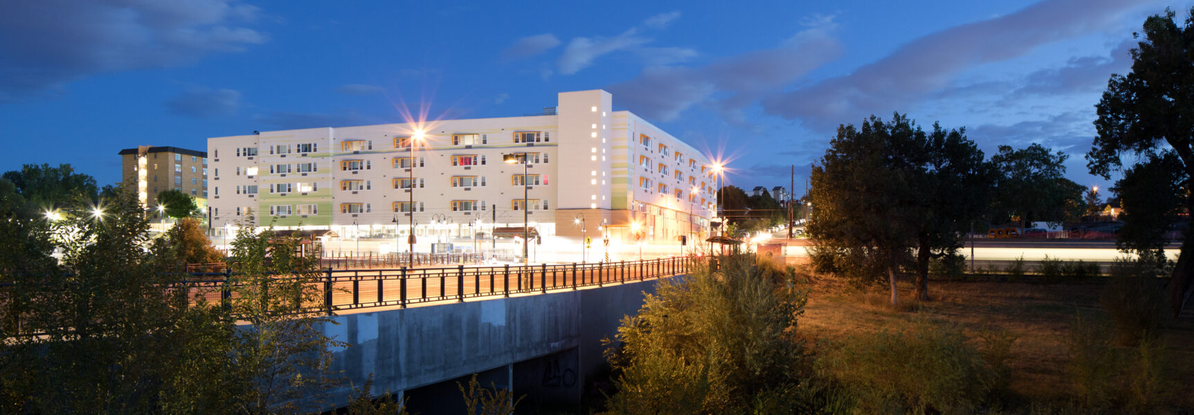 Illuminated multi-story building with balconies at dusk, surrounded by trees and a bridge in the foreground