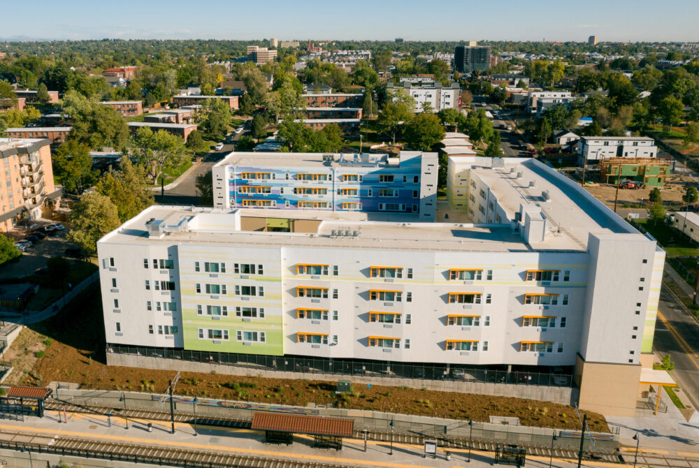Aerial view of a modern, multi-story apartment complex with colorful accents, surrounded by trees and residential neighborhood.