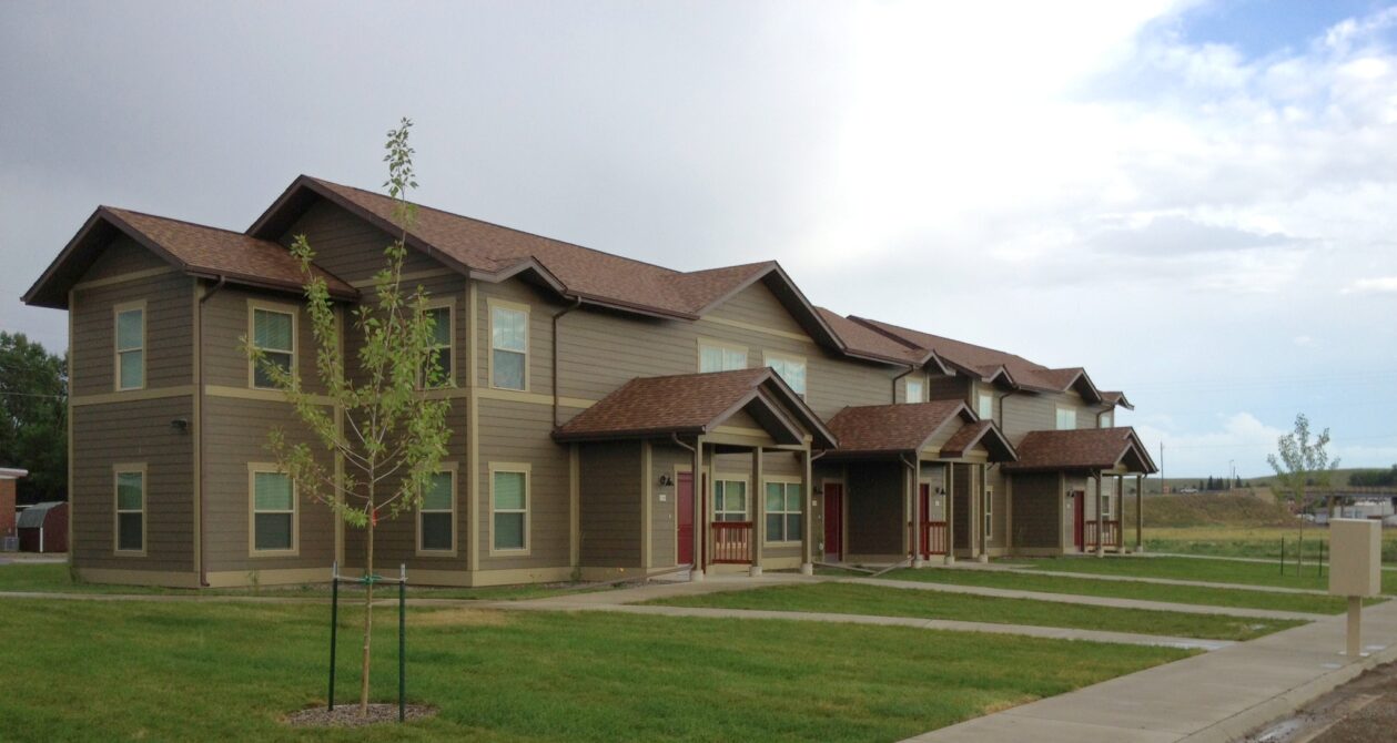 Brown modern apartment complex with multiple entrances, surrounded by green lawn and trees under a cloudy sky
