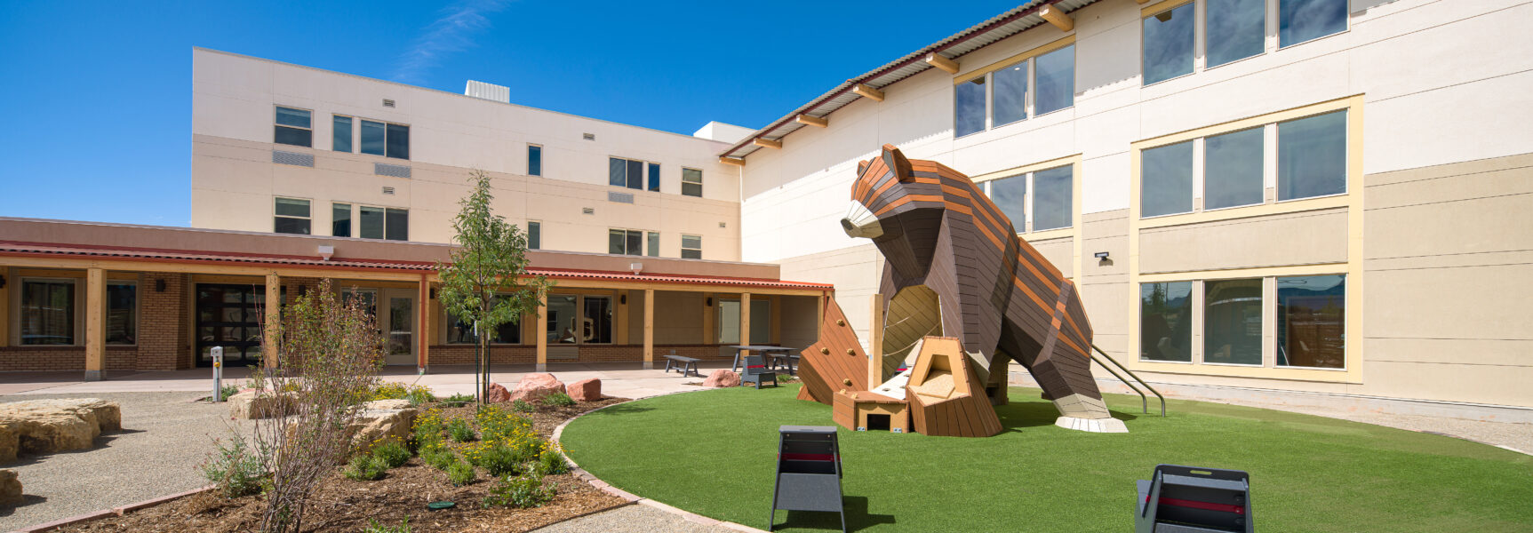 Courtyard with a playground featuring a large wooden bear sculpture, surrounded by greenery and modern buildings under a clear blue sky