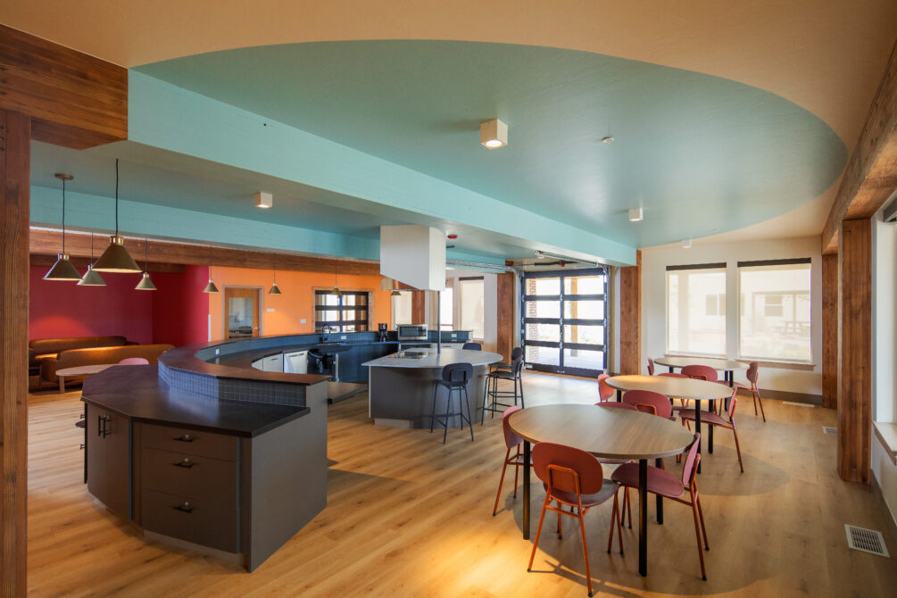 Modern open-plan kitchen and dining area with colorful walls, curved countertops, round tables, and large windows.