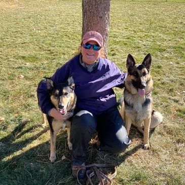 Tracy Lundgren smiles, sitting outdoors with two German Shepherds.