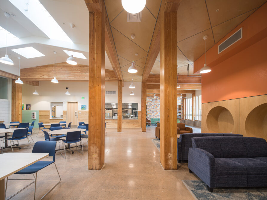 Spacious community room with wooden beams, tables, blue chairs, and sofas.