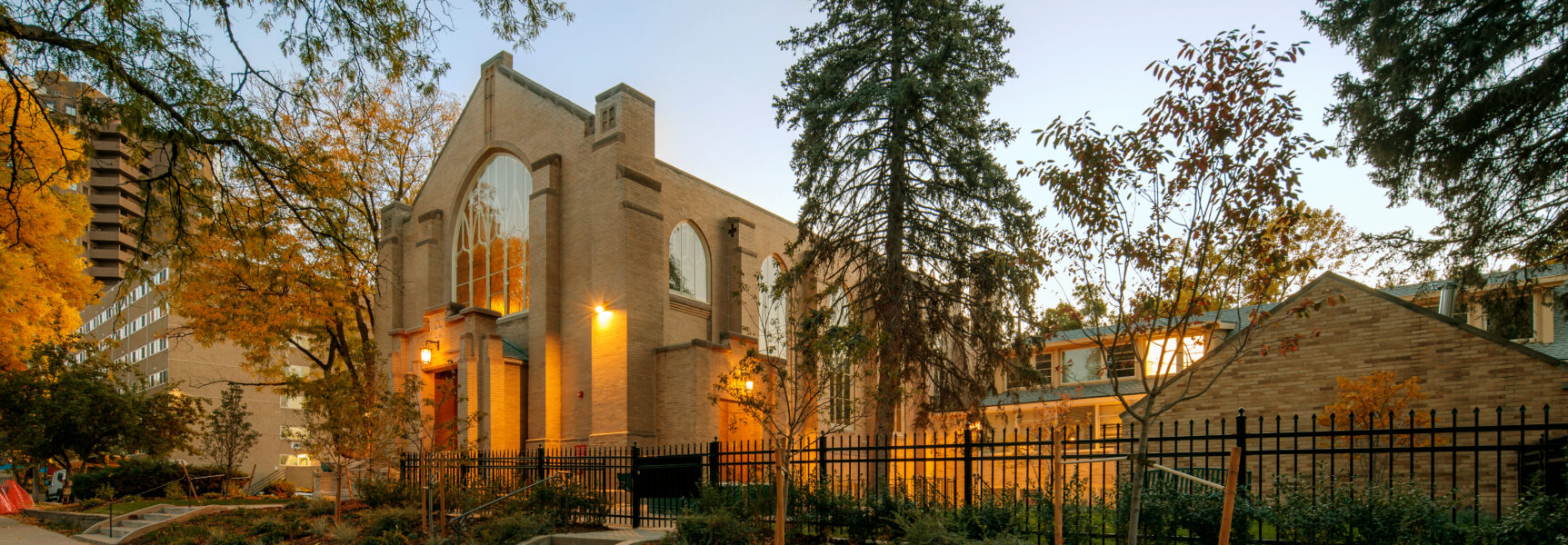Brick church building with large arched windows, surrounded by autumn trees and iron fencing