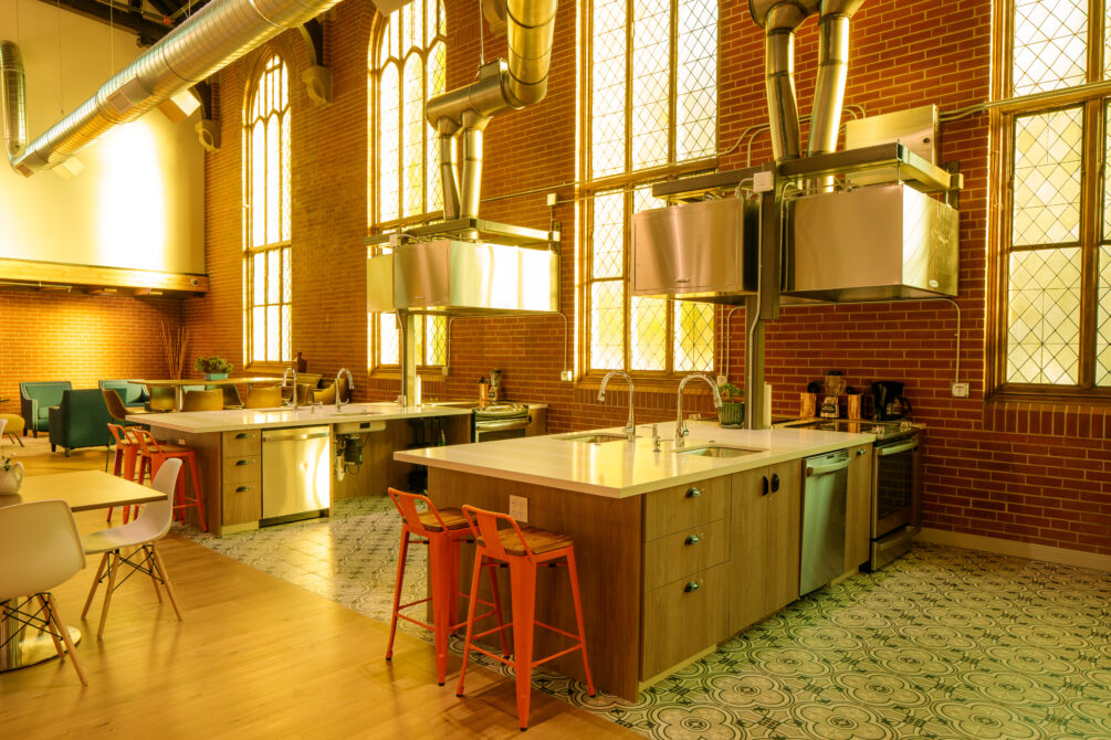 Spacious communal kitchen with high ceilings, large windows, brick walls, and two central islands.