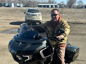 Clay sitting on a black motorcycle, smiling outdoors in a rural area.