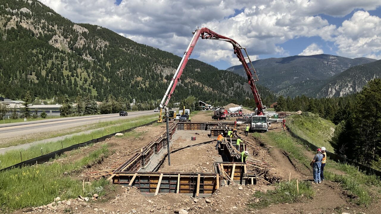 Construction workers pouring concrete into a foundation with mountainous landscape in the background.