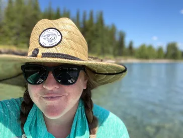 Marisa wearing sunglasses and a straw hat, smiling by a clear lake with trees in the background