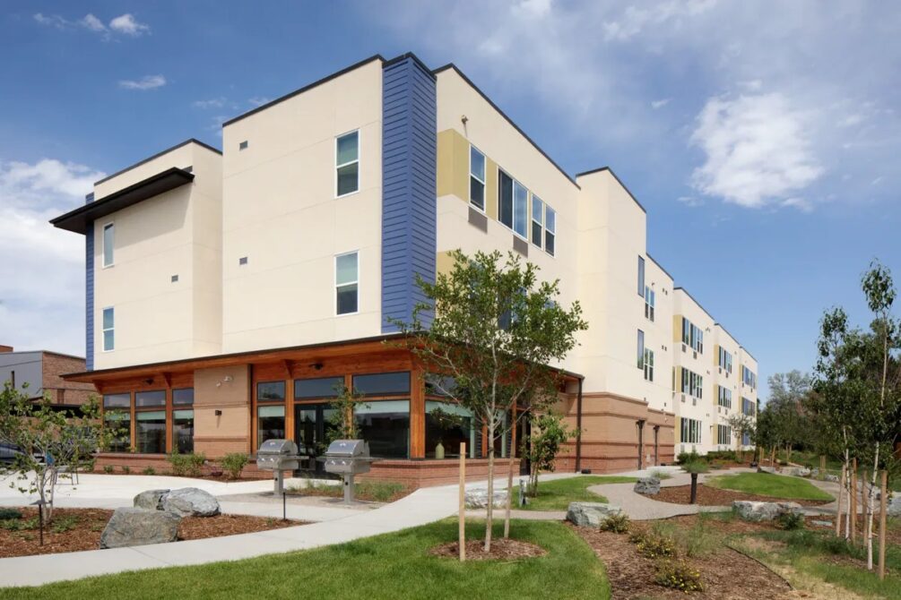 Modern three-story apartment building with landscaping and sidewalk under a blue sky