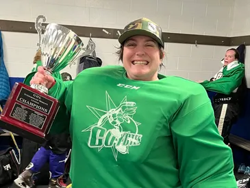Sarah, in a green hockey uniform, smiling and holding a championship trophy in a locker room.
