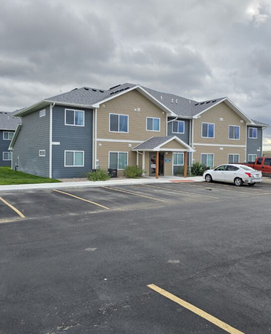 Apartment building with tan and blue siding under overcast sky, surrounded by empty parking lot.
